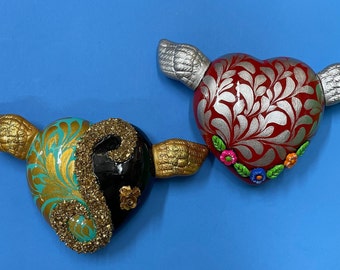 Könnte beinhalten: Zwei Keramik-Herzornamente mit Flügeln. Ein Herz ist rot mit silbernen Blumenmustern und bunten Blumen. Das andere Herz ist schwarz mit goldenen und türkisfarbenen Mustern und Glitzer.