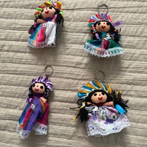 May include: Four doll keychains with black yarn hair, painted faces, and colorful ribbon headpieces. Each doll wears a fabric dress with lace trim and a small white tag numbered 43, 44, 45, or 46. The dolls feature varied patterns and colors.
