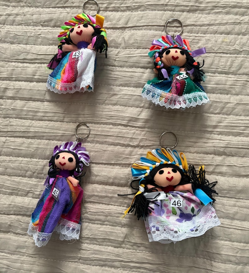May include: Four doll keychains with black yarn hair, painted faces, and colorful ribbon headpieces. Each doll wears a fabric dress with lace trim and a small white tag numbered 43, 44, 45, or 46. The dolls feature varied patterns and colors.
