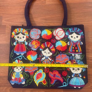 May include: A dark blue tote bag featuring colorful embroidered figures and floral patterns. A yellow measuring tape is placed across the center, showing a width of approximately 19 inches. The bag has two dark blue handles and a zipper closure.