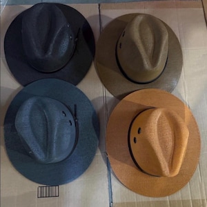 Puede incluir: Cuatro sombreros de paja en diferentes colores: negro, marrón claro, verde azulado y naranja. Cada sombrero tiene una copa redondeada, un ala ancha y una banda negra alrededor de la base de la copa. Los sombreros están colocados sobre una superficie de cartón.