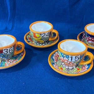 May include: Four ceramic espresso cups and matching saucers featuring vibrant, multi-colored floral patterns with orange rims. Each cup is labeled with a white sticker displaying the numbers 36, 24, 15, and 41, respectively, on the front.