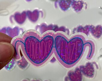 May include: A sticker featuring heart-shaped sunglasses with the text "Lavender Haze." The sunglasses are primarily purple with a glittery effect and a blue outline. Other similar stickers are scattered around.