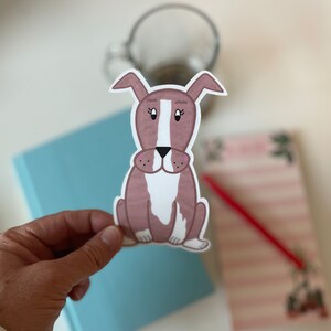May include: A sticker of a cartoon dog with brown and white fur. The dog has a white stripe down its face and pink nose. The dog is sitting with its legs crossed.