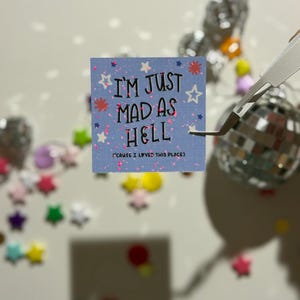 May include: A square, light blue card with the text "I'M JUST MAD AS HELL" and the phrase "(CAUSE I LOVED THIS PLACE)". The card is decorated with white stars and pink glitter. A pair of tweezers holds the card.