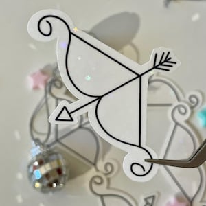 May include: A white sticker with a black outline of an arrow and bow. The sticker has a glittery finish.