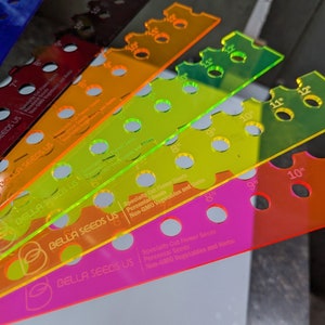 May include: Six colorful plastic rulers with holes for planting seeds. The rulers are labeled with inches and the text "BELLA SEEDS US". The rulers are for specialty cut flower seeds, perennial seeds, and non-GMO vegetables and herbs.