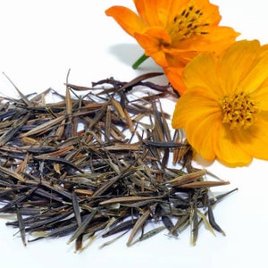 May include: A pile of dried brown and black cosmos flower seeds on a white background. Two orange cosmos flowers are in the background.