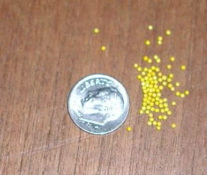May include: A silver dime next to a small pile of yellow beads on a brown wooden surface. The dime shows a profile of a person and the words "Liberty" and "United States of America". The beads are small and round.