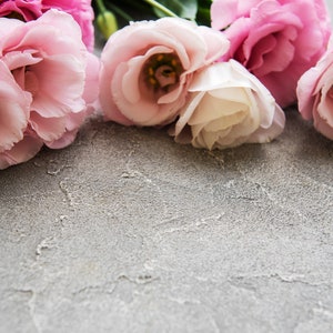 May include: A collection of pink and white lisianthus flowers with green stems and buds, arranged on a textured gray surface. The flowers have multiple layers of delicate petals.