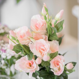 May include: A bouquet of soft pink roses with green leaves and buds. The flowers are in full bloom, with delicate petals and a natural appearance. The arrangement is set against a blurred background, suggesting a light and airy setting.