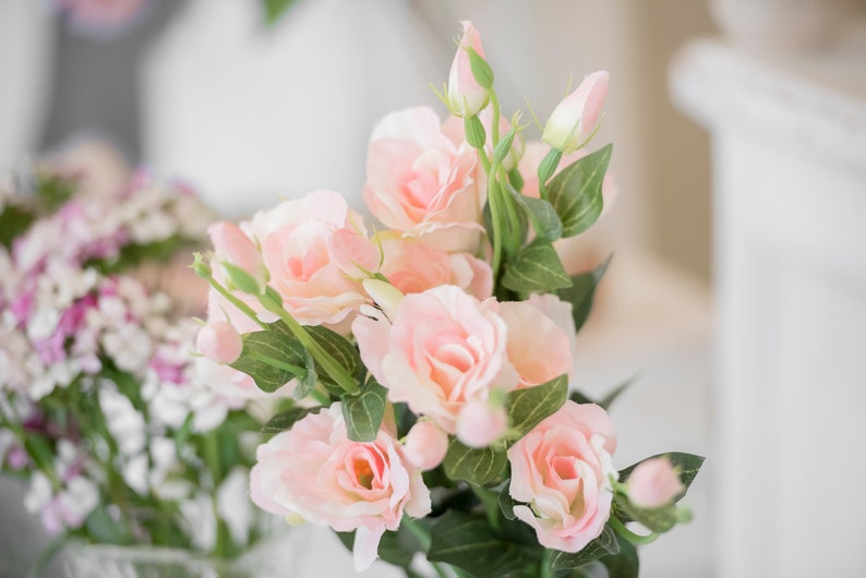 May include: A bouquet of soft pink roses with green leaves and buds. The flowers are in full bloom, with delicate petals and a natural appearance. The arrangement is set against a blurred background, suggesting a light and airy setting.