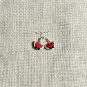 May include: A pair of watermelon slice earrings. Each earring features a red watermelon slice with black seeds and a white rind. The earrings are attached to silver-colored fish hook ear wires. The earrings are set against a neutral background.