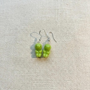 May include: A pair of lime green gummy bear earrings. Each earring features a small, stylized gummy bear charm with visible facial features and a rounded body. The charms hang from silver-colored fish hook ear wires.