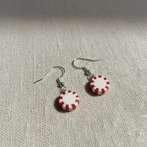 May include: A pair of dangle earrings featuring a peppermint candy design. Each earring has a white base with red stripes around the edge. The earrings have silver-colored hooks.