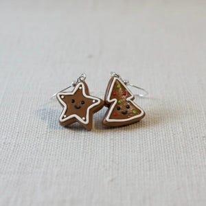 May include: A pair of festive earrings featuring a star and a Christmas tree design. The earrings are brown with white icing details and small, colorful accents. Each earring has a smiling face.