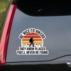 May include: A sticker with a hiker graphic and the text "BE NICE TO HIKERS THEY KNOW PLACES YOU'LL NEVER BE FOUND." The design features a hiker silhouette, mountains, and a sunset in shades of orange and yellow.