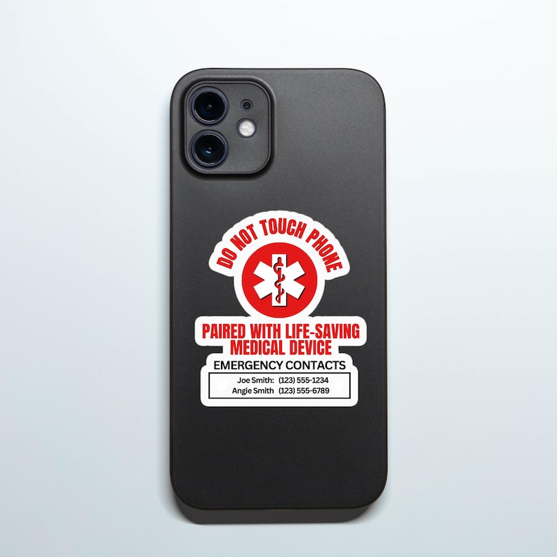Op de afbeelding: Zwarte telefoonhoes met een rood-witte sticker. De sticker luidt "NIET AANRAKEN TELEFOON" en "GEPAARD MET LEVENSREDDEND MEDISCH APPARAAT". Daaronder staan ​​noodcontacten met namen en telefoonnummers. De telefoonhoes heeft een dubbele cameralens.