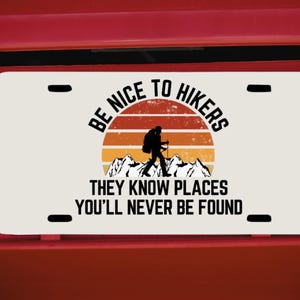 May include: A white license plate with the text "BE NICE TO HIKERS THEY KNOW PLACES YOU'LL NEVER BE FOUND." The design features a hiker silhouette against a sunset and mountain range.