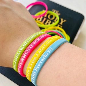 May include: A wrist with several colorful silicone bracelets with white lettering. The bracelets have bible verses printed on them. The bracelets are yellow, pink, light blue, and green.