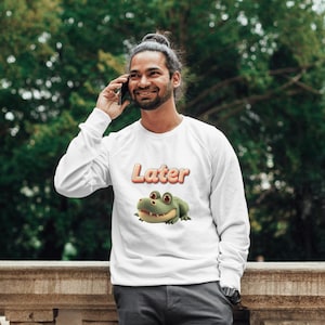 Peut inclure: Sweat-shirt blanc à manches longues avec le mot "Later" en orange et un dessin de grenouille. La grenouille est verte avec des yeux rouges. Le sweat-shirt est porté par une personne.