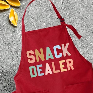 May include: Red kitchen apron with the text "SNACK DEALER" in colorful letters. The apron has two pockets and adjustable straps.