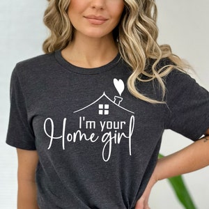 May include: Dark gray t-shirt with the text "I'm your home girl" in a white script font. The design includes a house silhouette with a heart-shaped smoke plume. The shirt is a short-sleeved crew neck.