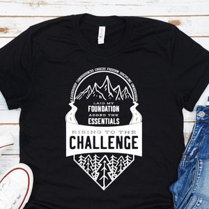 May include: Black t-shirt with a white graphic design featuring a mountain range and the text "Laid my foundation, added the essentials, Rising to the challenge".