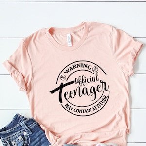 Peut inclure: Un t-shirt rose avec un graphique circulaire noir qui dit "Avertissement&nbsp;: Adolescent officiel Peut contenir de l'attitude".