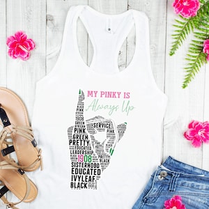 May include: A white racerback tank top featuring a graphic of a hand gesture formed by words including 1908, sisterhood, and leadership. The text My Pinky Is Always Up is printed in pink and green. The design is centered on the front of the garment.
