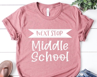 May include: A pink t-shirt with the text "Next Stop Middle School" in white. The shirt is laid flat on a white wooden surface with a pair of brown sandals and blue jeans.