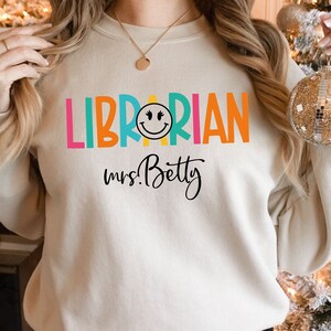 May include: A white sweatshirt with the text "LIBRARIAN" in rainbow colors and a smiley face. The text "mrs. Betty" is written in black below the word "LIBRARIAN".