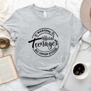 Peut inclure: T-shirt gris avec un graphique circulaire noir qui dit "Avertissement&nbsp;: Adolescent officiel Peut contenir de l'attitude".