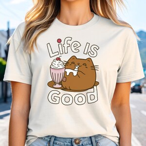 May include: A white t-shirt with a brown cat cartoon illustration drinking a milkshake with a cherry on top. The text "Life is Good" is printed in black with a dotted outline.