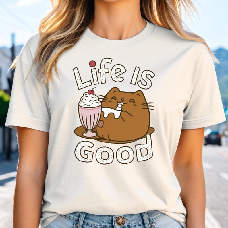 May include: A white t-shirt with a brown cat cartoon illustration drinking a milkshake with a cherry on top. The text "Life is Good" is printed in black with a dotted outline.