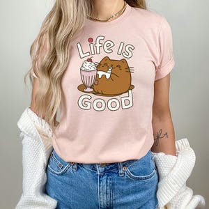 May include: A pink t-shirt with a cartoon cat drinking a milkshake. The text "Life is Good" is printed on the shirt.
