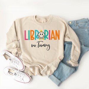May include: A beige sweatshirt with the text "LIBRARIAN" in rainbow colors and a smiley face. The text "Mr. Tommy" is written in black below the word "LIBRARIAN".