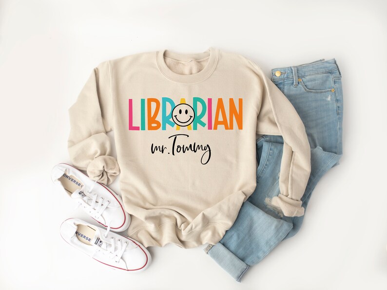 May include: A beige sweatshirt with the text "LIBRARIAN" in rainbow colors and a smiley face. The text "Mr. Tommy" is written in black below the word "LIBRARIAN".