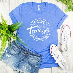 Peut inclure: Un t-shirt bleu avec un graphique circulaire blanc qui dit "Avertissement&nbsp;: Adolescent officiel Peut contenir de l&rsquo;attitude". Le t-shirt est pos&eacute; sur une surface en bois blanc avec une paire de baskets blanches et un short en jean.