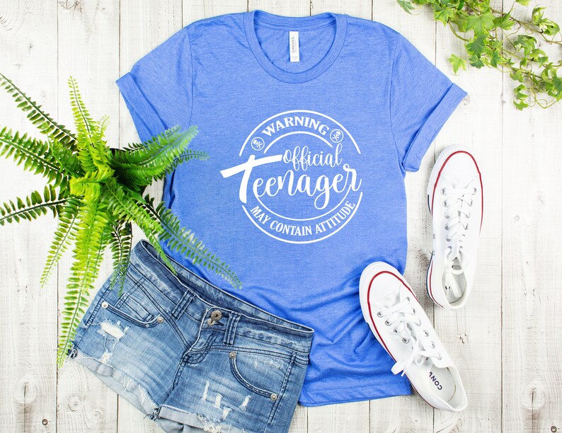 Peut inclure: Un t-shirt bleu avec un graphique circulaire blanc qui dit "Avertissement&nbsp;: Adolescent officiel Peut contenir de l&rsquo;attitude". Le t-shirt est pos&eacute; sur une surface en bois blanc avec une paire de baskets blanches et un short en jean.