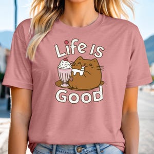 May include: A pink t-shirt with a cartoon cat enjoying a milkshake. The text "Life is Good" is printed on the shirt.
