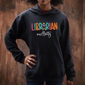 May include: Black hooded sweatshirt with the text "LIBRARIAN" in rainbow colors and a smiley face. The text "mrs. Betty" is written below in white.