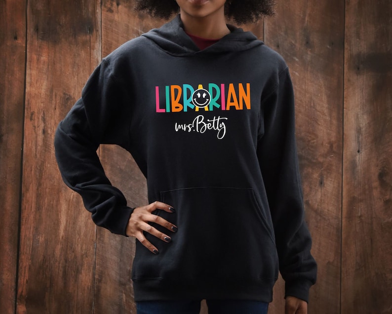 May include: Black hooded sweatshirt with the text "LIBRARIAN" in rainbow colors and a smiley face. The text "mrs. Betty" is written below in white.