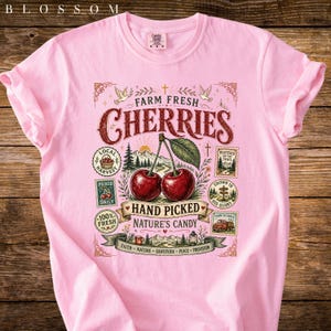 May include: A light pink t-shirt featuring a graphic of two cherries with the text "Farm Fresh Cherries". The design includes additional text such as "Hand Picked" and "Nature's Candy".
