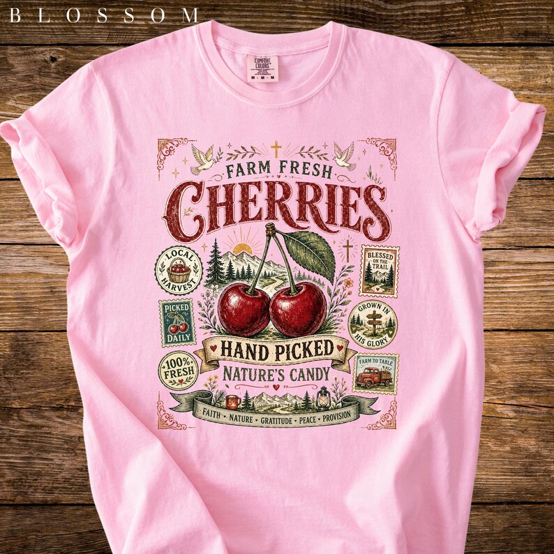 May include: A light pink t-shirt featuring a graphic of two cherries with the text "Farm Fresh Cherries". The design includes additional text such as "Hand Picked" and "Nature's Candy".