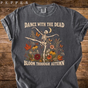 May include: A charcoal gray crew neck t-shirt featuring a graphic of a skeleton dancing amidst autumn flowers, leaves, butterflies, and a crescent moon. The text reads DANCE WITH THE DEAD and BLOOM THROUGH AUTUMN in a light tan, vintage-style font.