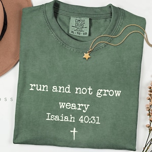 May include: An olive green t-shirt with the text "run and not grow weary Isaiah 40:31" in white. A gold star pendant necklace rests on the shirt. The shirt has a Comfort Colors tag, size XL.
