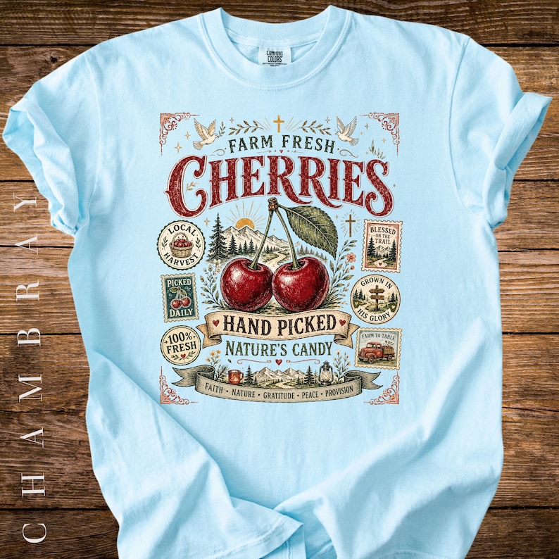 May include: Light blue t-shirt with a graphic featuring the words "Farm Fresh Cherries" and an illustration of two cherries. The design includes additional text and imagery related to farming and nature.