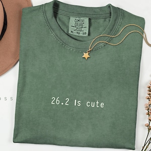 Op de afbeelding: Een saliegroen t-shirt met de tekst "26.2 is cute" in het wit. Het shirt heeft een ronde hals en korte mouwen. Een gouden sterhangerketting ligt op het shirt. Het shirt is maat XL.