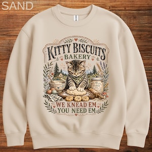 May include: A sand-colored crewneck sweatshirt featuring a graphic of a tabby cat kneading dough. The text reads KITTY BISCUITS BAKERY and WE KNEAD EM YOU NEED EM. The illustration includes a flour jar, a bowl, eggs, and botanical elements.